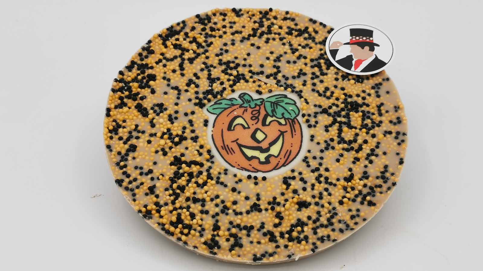 Schokomichi Scheibe aus weisser Schokolade mit einer Dekormasse mit Kürbismotiv in der Mitte und flächig, mit selbst gemischten Halloween-Streussel in Orange und Schwarz, bestreut.