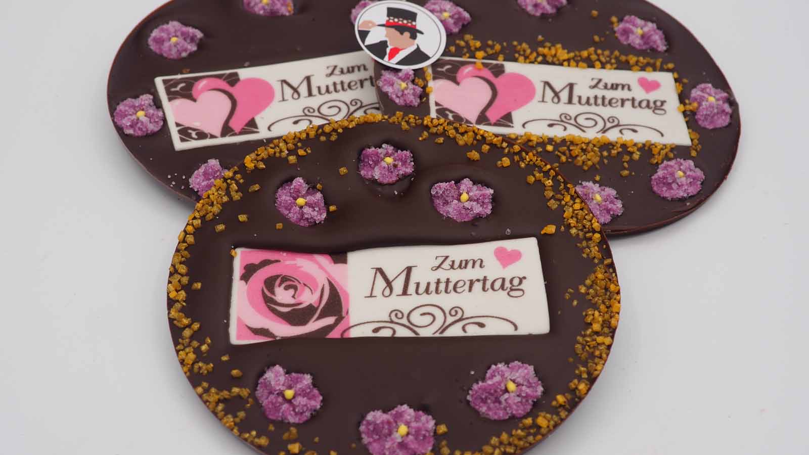 Handgezogene Scheiben aus Zartbitterschokolade mit Zuckerblümchen, Flimmerzuckerrand und Dekormasse (rosa Herzen oder Rose, schokobraune Verzierung, Schriftzug: »Zum Muttertag«) zum Muttertag.