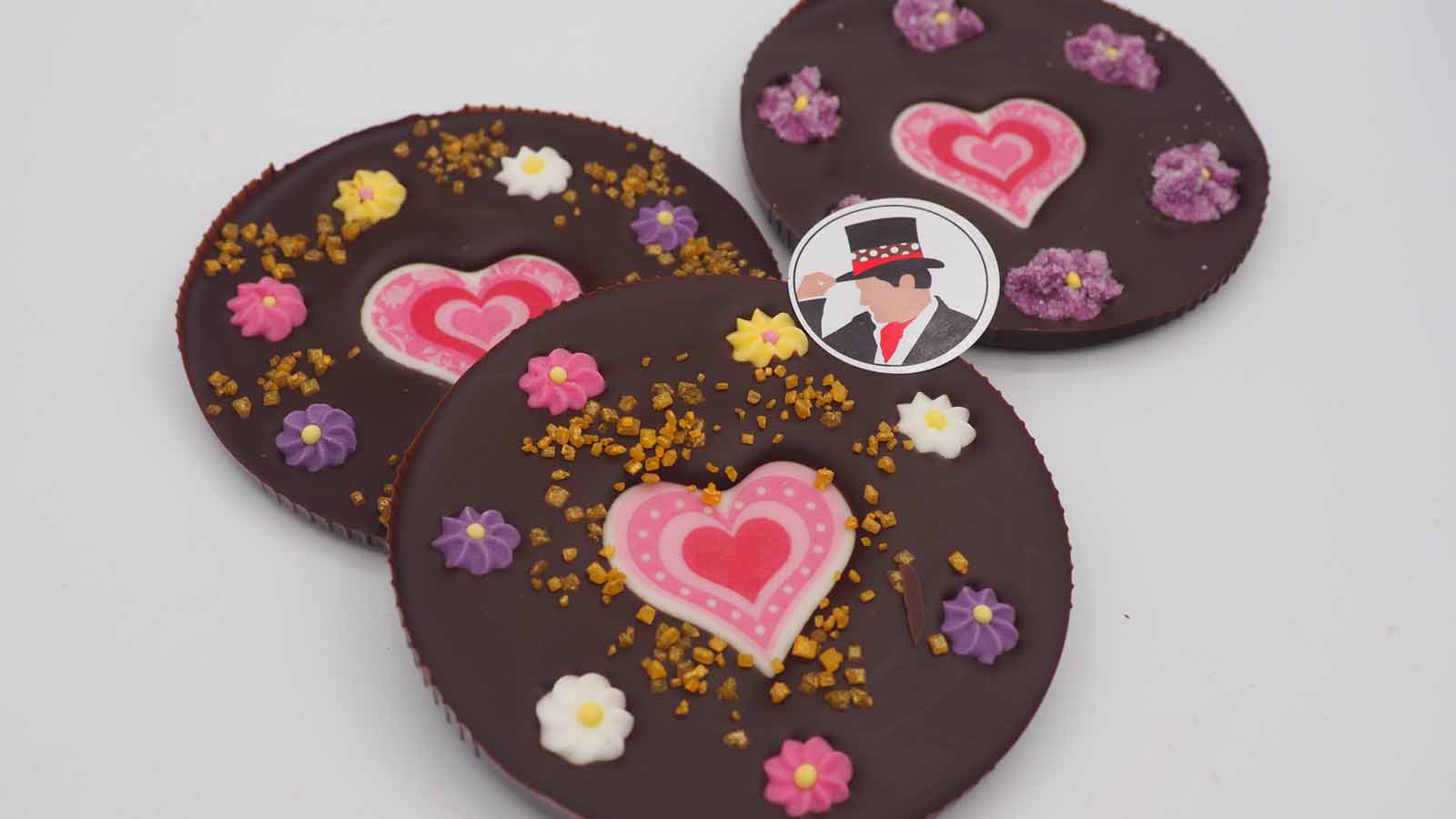 Schokomichi Scheiben aus feiner Zartbitterschokolade mit Zuckerherzen und Zuckerblümchen zum Muttertag Handgezogene Scheiben aus Zartbitterschokolade mit Zuckerherzen, Zuckerblümchen und Dekormasse (rosa Herzen in verschiedenen Variationen) zum Muttertag.