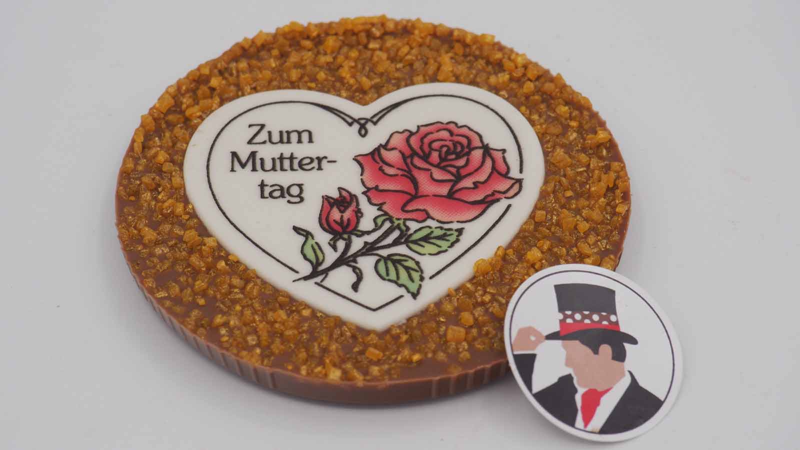 Handgezogene Scheibe aus Vollmilchschokolade mit viel Flimmerzucker und Dekormasse (rosa Rose, Schriftzug: »Zum Muttertag«) zum Muttertag.