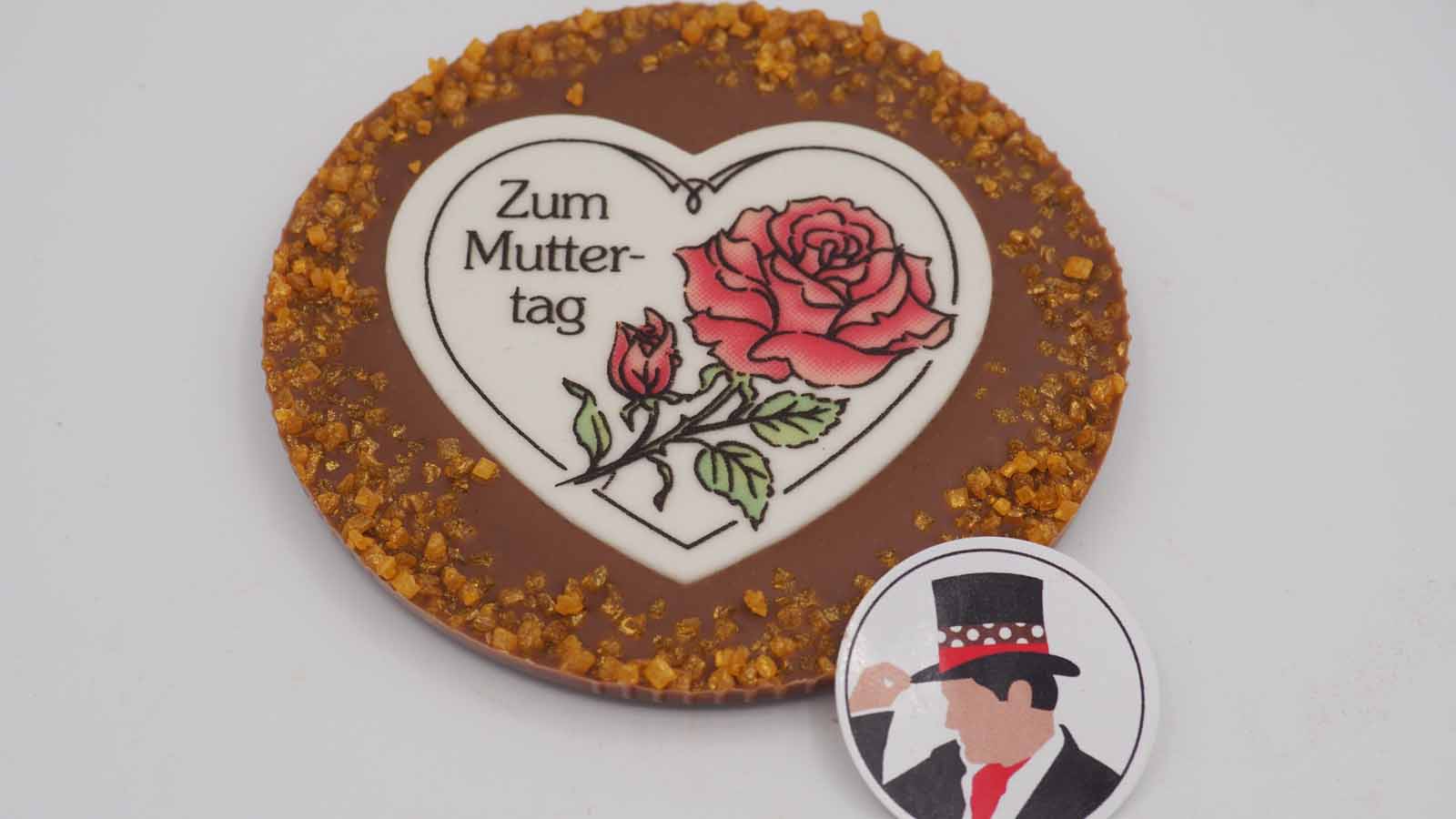 Handgezogene Scheibe aus Vollmilchschokolade mit Flimmerzuckerrand und Dekormasse (rosa Rose, Schriftzug: »Zum Muttertag«) zum Muttertag.