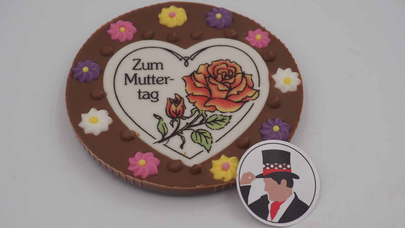 Handgezogene Scheibe aus Vollmilchschokolade mit Zuckerblümchen und Dekormasse (rosa Rose, Schriftzug: »Zum Muttertag«) zum Muttertag.