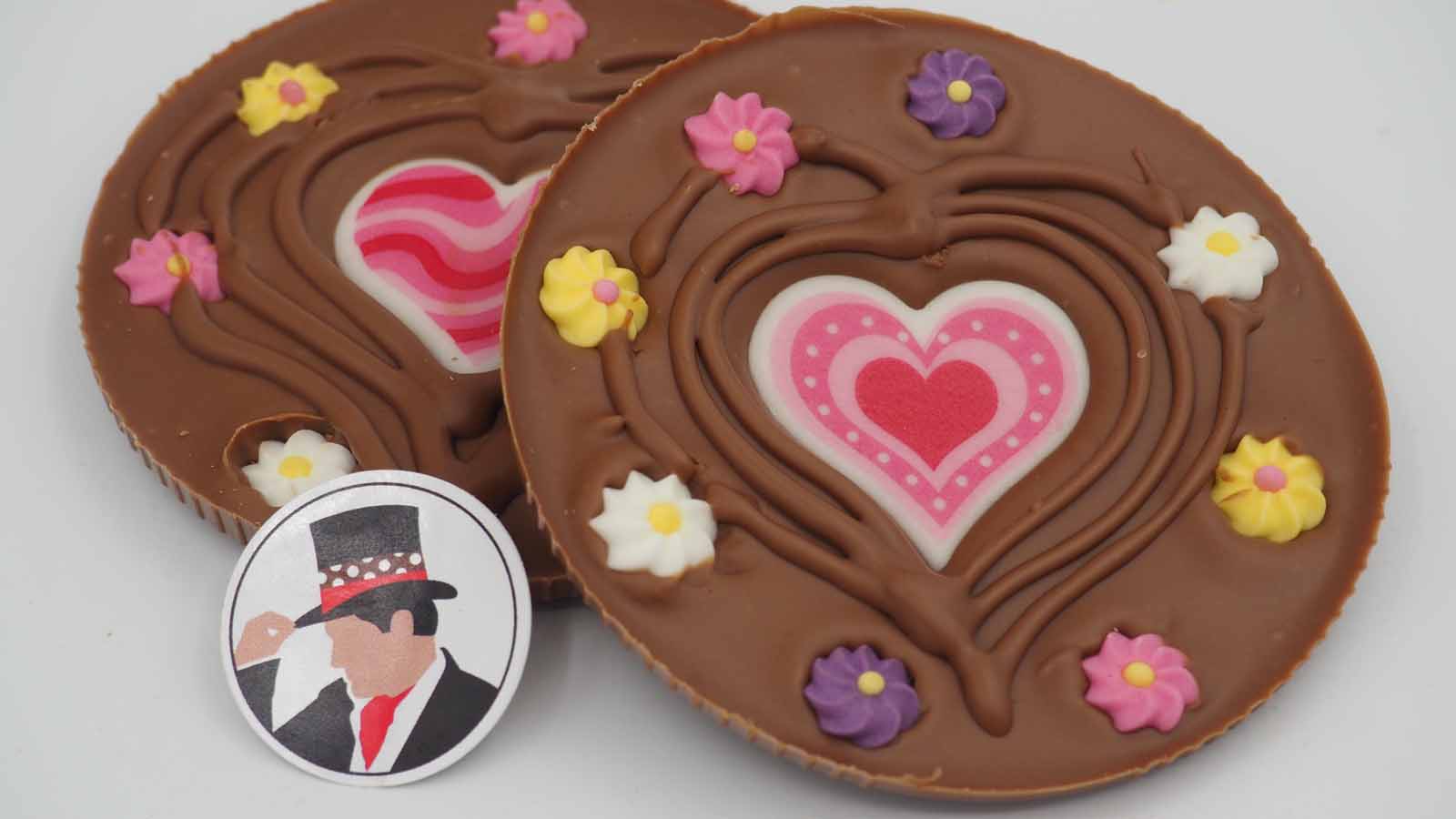 Schokomichi Scheibe aus Vollmilchschokolade mit Dekorherz und Zuckerblümchen zum Muttertag Handgezogene Scheibe aus Vollmilchschokolade mit Dekormasse (rosa in verschiedenen Variationen) und Zuckerblümchen zum Muttertag.