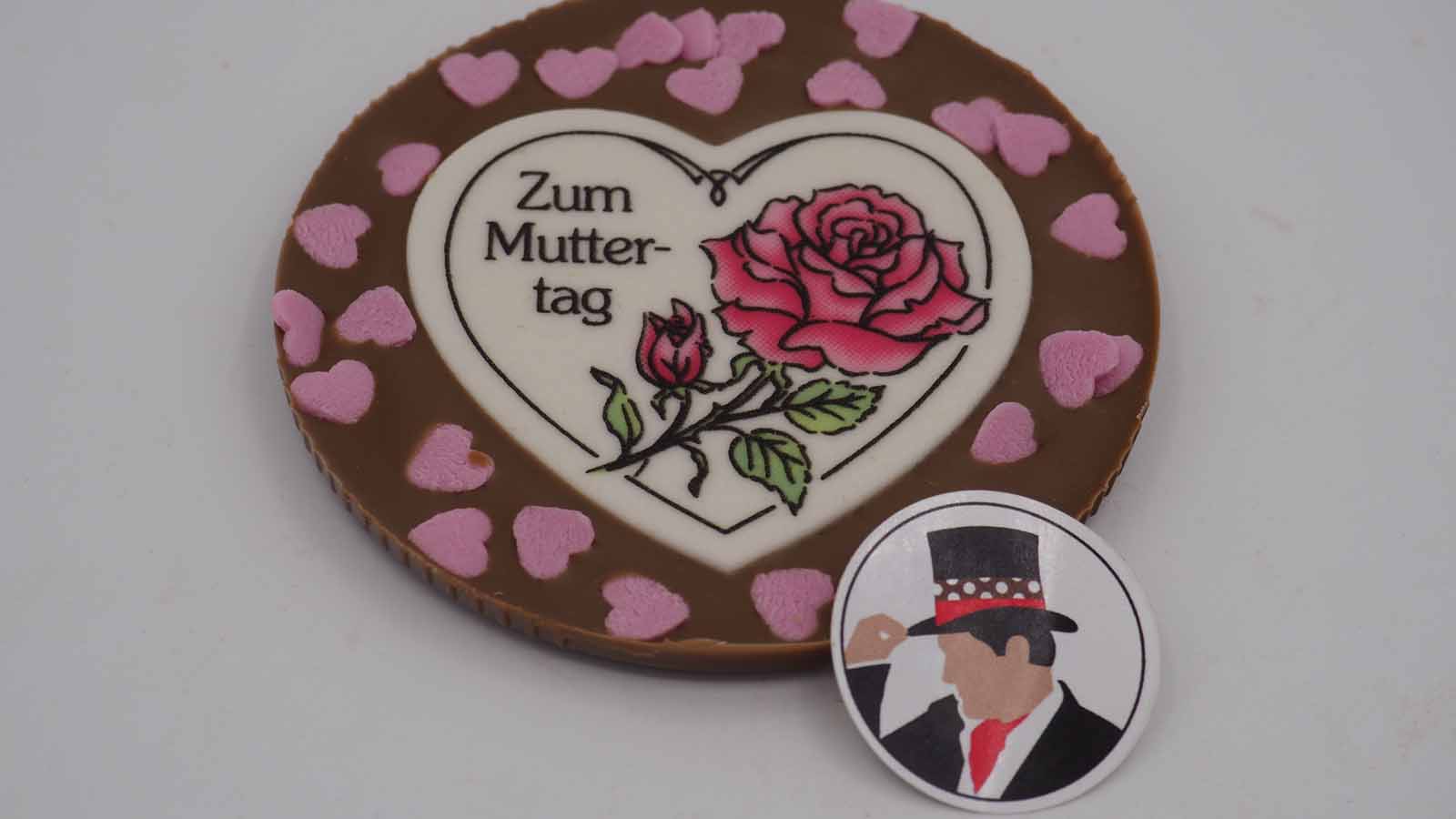 Schokomichi Scheibe aus Vollmilchschokolade mit Dekormasse und rosa Zuckerherzen zum Muttertag Handgezogene Scheibe aus Vollmilchschokolade mit Dekormasse (rosa Rose, Schriftzug: »Zum Muttertag«) und rosa Zuckerherzen zum Muttertag.