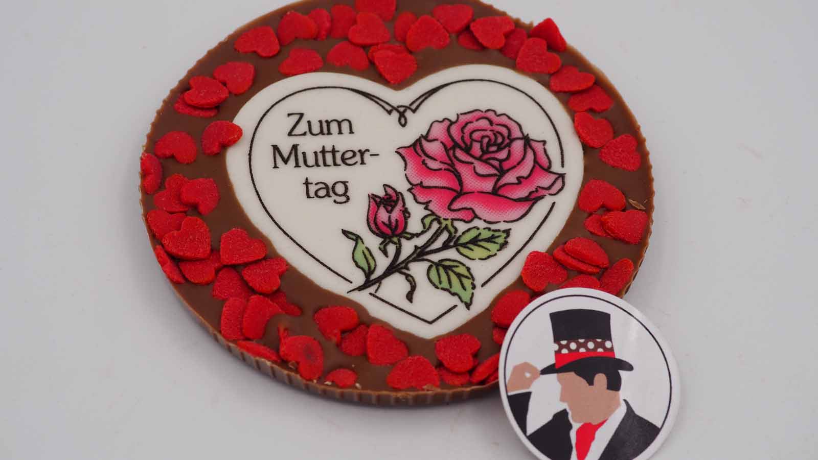 Handgezogene Scheibe aus Vollmilchschokolade mit Dekormasse (rosa Rose, Schriftzug: »Zum Muttertag«) und roten Zuckerherzen zum Muttertag.
