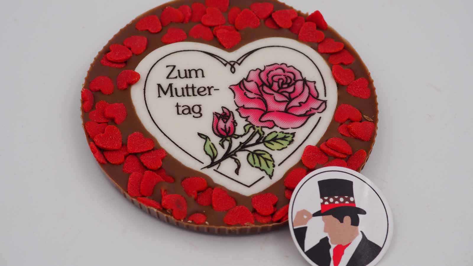 Schokomichi Scheibe aus Vollmilchschokolade mit roten Zuckerherzen zum Muttertag Handgezogene Scheibe aus Vollmilchschokolade mit Dekormasse (rosa Rose, Schriftzug: »Zum Muttertag«) und roten Zuckerherzen zum Muttertag.