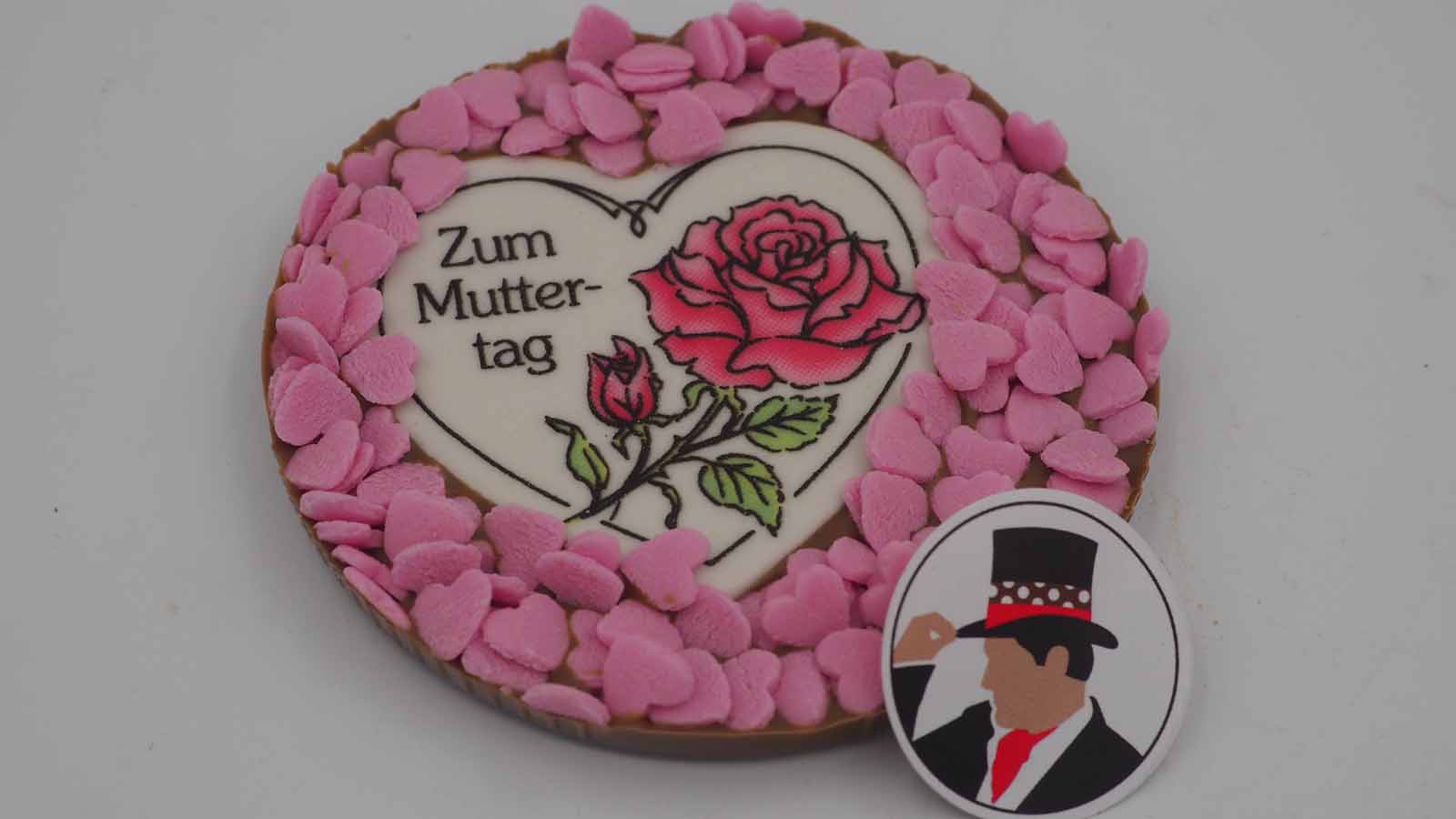 Handgezogene Scheibe aus Vollmilchschokolade mit Dekormasse (rosa Rose, Schriftzug: »Zum Muttertag«) und vielen rosa Zuckerherzen zum Muttertag.