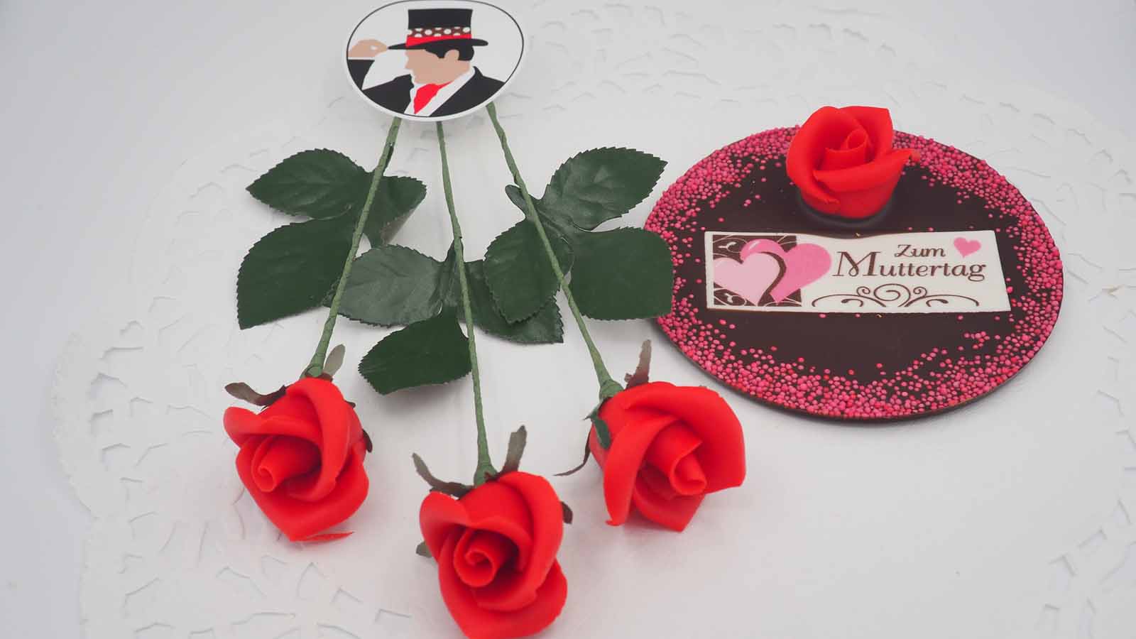 Schokomichi Marzipanrosen am Stiel und eingestreut in einer Scheibe aus Zartbitterschokolade Rosenstrauß aus handgemachten Marzipanrosen am Stiel und eine handgezogene Scheibe aus Zartbitterschokolade, bestreut mit einer Marzipanrose, Streussel und Dekormasse (rosa Herzen, schokobraune Verzierung, Schriftzug: »Zum Muttertag«) zum Muttertag.