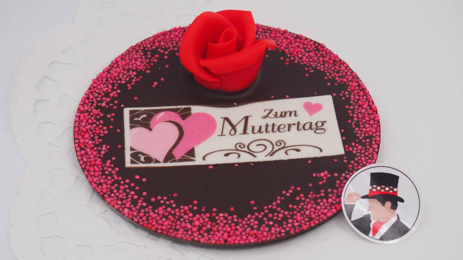 Schokomichi Marzipanrose eingestreut in einer Scheibe aus Zartbitterschokolade mit rosa Streussel Handgezogene Scheibe aus Zartbitterschokolade, bestreut mit einer handgemachten Marzipanrose, rosa Streussel und Dekormasse (rosa Herzen, schokobraune Verzierung, Schriftzug: »Zum Muttertag«) zum Muttertag.